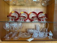 Full view of shelf showing reddish plates and glasses with crystal bowls, salt dispensers, tray, and pitcher on wood-backed shelf