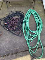 Three hoses laid out on concrete; green garden hose and two black nylon-coated hoses with a purple expandable hose looped on top.