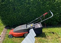 Side view of the Black & Decker orange and black electric mulching lawn mower with attached grass catcher and power cord, sitting on grass in front of a hedge.