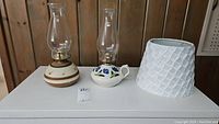 Two oil lamps and one white lamp shade on a white surface against a wooden background. Lamps have clear glass chimneys with ceramic bases, one decorated with blue flowers and the other with brown stripes. The lamp shade is white with a textured pattern.