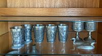 Five pewter cups, two pewter goblets, and two metal duck figurines arranged on a glass shelf with wooden backing. The pewter items have detailed embossed designs with scenic elements.