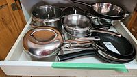 Drawer showing various stainless steel pots, pans, and colanders with handles and lids.