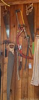 Photo showing various saws mounted on a wooden wall alongside a red crowbar and an axe with wooden handle