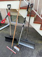 Full view of five outdoor tools: two shovels for snow removal, a soft scrub brush, a squeegee, and a plastic outdoor brush leaning against stairs outdoors.
