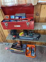 Photo of the red metal toolbox closed with a view of additional hand tools on shelves below including clamps and saws.