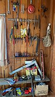 Photo showing a pegboard wall with various hanging tools including wrenches, screwdrivers, pliers, scissors, rope, and measuring tools on shelves below.