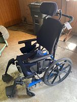 Side angled view of the full wheelchair showing blue frame, large wheels, black padded seat and neck rest, armrests, footrests, and rear handles.