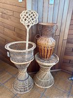 Photo showing two wicker plant stands side by side, with the wicker vase placed on the shorter stand, and a wicker fan standing in the taller stand.