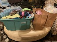 Green plastic bin filled with assorted yarn skeins and balls in various colors including blue, yellow, and brown, plus two brown paper bags beside the bin.