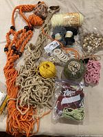 Photo of assorted macrame supplies and partial projects on a cloth surface showing braided cords, yarn balls, wooden beads, rings, and bags with craft materials.