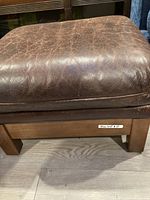 Close-up of brown leather ottoman showing wear and creased surface with sturdy wooden legs