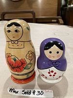 Two nesting dolls displayed side by side on a table, one wooden doll with floral painting, another smaller purple and white doll with painted cherry design.