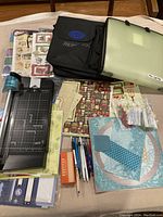 Lot laid out showing black rotary paper cutter with visible blade system and corner rounder, assorted patterned paper sheets, pens, staples, and storage cases.