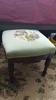 Full side view of antique wooden ottoman stool with needlepoint upholstered floral top, showing carved legs and casters.