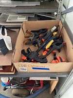 Cardboard box filled with assorted clamps including black and orange quick-grip clamps and wooden-handled clamps, placed on a shelf.