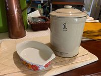 Vintage 1 gallon Medalta crock pot with lid and a small floral dish beside it on a table