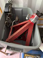 Two red Motomaster 6 ton axle stands and a metal hoist tool inside a large gray bin.