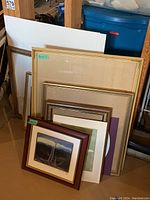 Set of frames of various sizes stacked together, some with glass and some without, placed in basement area.