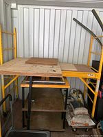 Wide angle showing full assembled scaffolding setup with yellow steel frames supporting plywood platforms and wood boards, bags of materials stored underneath.