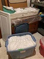 Blue plastic storage bins filled with white sheer curtains and drapery pins located in a basement space.