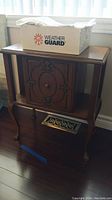 Wood side table cabinet with decorative front door and box of embroidered linens on top.
