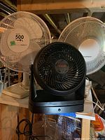 Four fans: three white and one black, all table top models with protective grilles and adjustable bases. Located on a shelf in a basement setting.