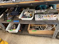 Metal shelf with various tools and hardware including boxed Genesis oscillating tool, white plastic bins with assorted hand tools, and a cardboard box with hardware.