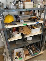 Metal shelving unit with various tools and hardware on shelves including hard hat, tool belt, and containers of small parts.