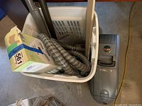 Photo showing the vacuum cleaner and accessories including hose, nozzles, and filter bags inside a white laundry basket on a concrete floor.