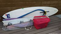 Overall view of the wind surfing board with mast, sail in carrying bag, and red container.