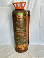 Full view of antique brass fire extinguisher with orange safety cage on top, brass discharge tube attached to side, and patinated surface.