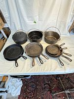 Wide shot showing the full lot of 13 antique cast iron pans arranged on white surface including Dutch ovens with bail handles and multiple skillets of various sizes.