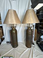 Two full view images of the pair of antique brass fire extinguisher lamps standing side by side, showing their beige lampshades and wooden table surface.