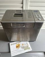 Front and top view of the Breadman breadmaker device showing stainless steel body, control panel with buttons and display, and user manual placed in front.