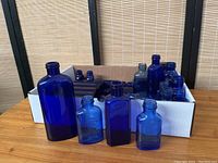Front view of several cobalt blue vintage bottles including large and smaller sizes displayed in front of a box containing additional bottles.