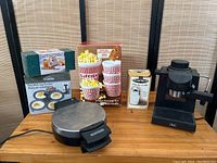 Photo of mixed kitchenware lot showing various boxed and unboxed items including popcorn bucket set, coffee maker, and metallic round appliance