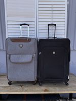 Front view of two large rolling suitcases side by side: grey Adolfo and black Travel Hardware