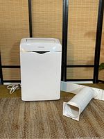 Front and side view of white NOMA portable air conditioner with attached white vent hose and window installation kit on a brown rug with bamboo background.