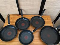 Overview of all five black T-Fal frying pans with long handles placed on wooden surface showing various sizes.