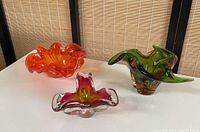 Three colorful blown glass bowls displayed on a white surface with a bamboo backdrop, showing overall shapes and colors.
