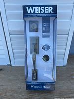Full view of the Weiser lock set in sealed retail packaging showing door handle, deadbolt, and keyhole with branding and security info visible.