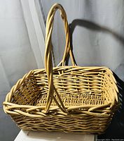 Photo showing large rectangular wicker basket made of light-colored woven wicker with a tall handle.