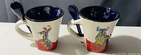 Two mini ceramic cups with colorful dancing figures and matching spoons on a white surface.