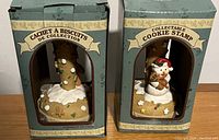 Photo showing two cookie stamps in their original packaging. One is a gingerbread man figure with decorative icing and candy, the other is a snowman figure holding a cookie. Both are mounted on cream-colored stamp bases inside green collectible boxes with windows.