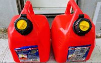 Photo shows a pair of red plastic gas cans with yellow and black safety caps. Labels indicate each can has a 5-gallon capacity. Both cans appear in good condition and ready for use.