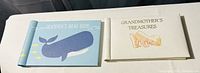 Photo of two small photo albums; left is light blue with a whale and yellow fish and text 'Grandma's Brag Book'; right is beige with text 'Grandmother's Treasures' and a baby shoes illustration.