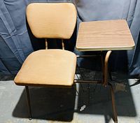 Front angled view of the whole Cooey table and chair combo, showing the brown vinyl padded chair and attached woodgrain table top against a dark backdrop.