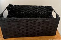 Photo of a rectangular black woven storage basket with side handles, placed on the floor against a white wall.