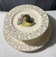 View of the stacked two-tier appetizer plates showing the detailed floral artwork in the center with gold-tone floral borders and brass knob handle on top tier plate.