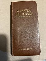 Front cover of the Webster Dictionary Self Pronouncing De Luxe Edition pocketbook, showing brown leather-like cover with embossed gold text.